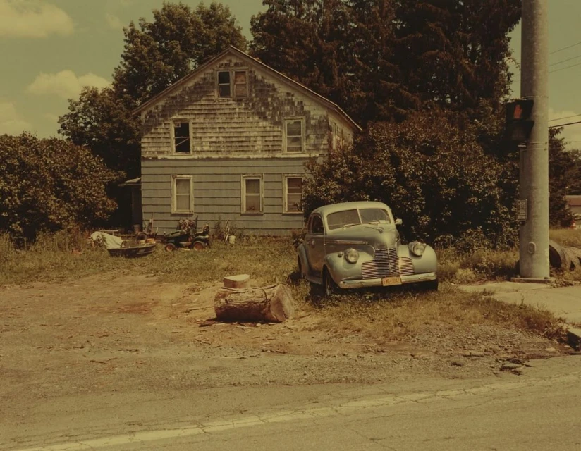 Vintage teal car parked on a grassy patch with a weathered two-story house in the background. Trees and sky visible.