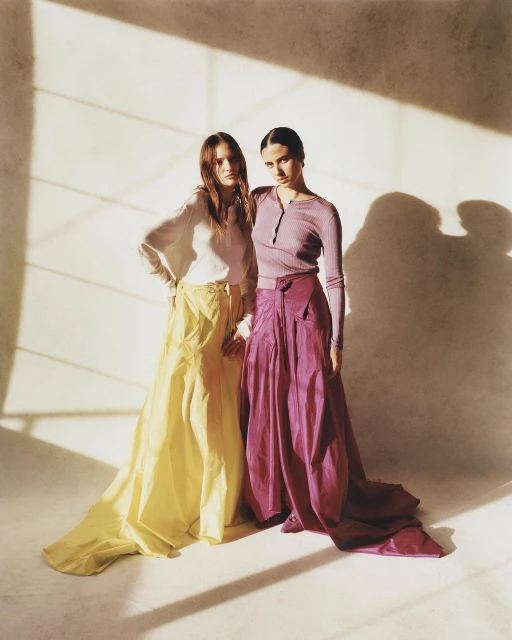 Two women pose indoors, wearing long skirts and long-sleeved tops; light shines through a window onto the models and background.