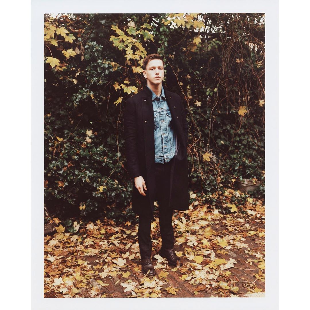 Full-length portrait of a young adult male in denim shirt and dark overcoat, standing outside on leaves in front of dense foliage.