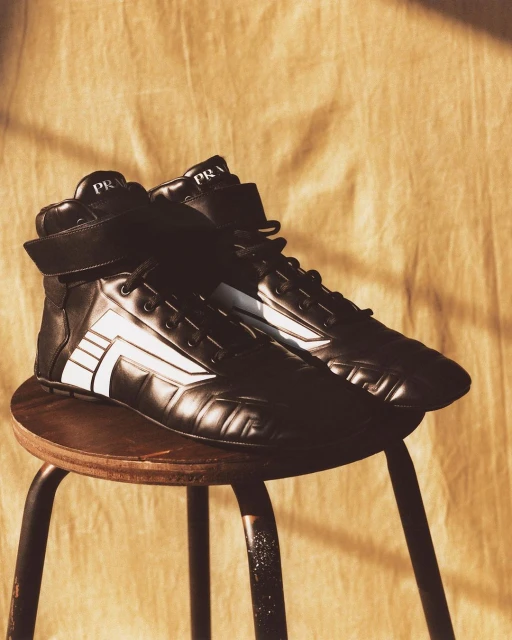 Prada high-top sneakers on a wooden stool against a textured background. The black shoes feature a white graphic design.