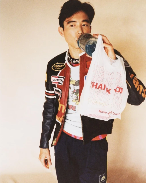 Asian man drinks from a clear bottle while holding a plastic shopping bag, wearing a racing jacket and athletic pants in a studio.