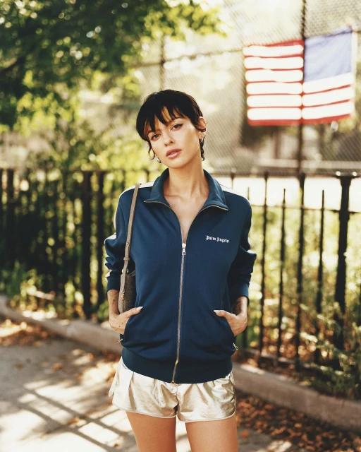 A woman poses in a teal jacket and gold shorts against a park background, featuring a glimpse of an American flag in the distance.