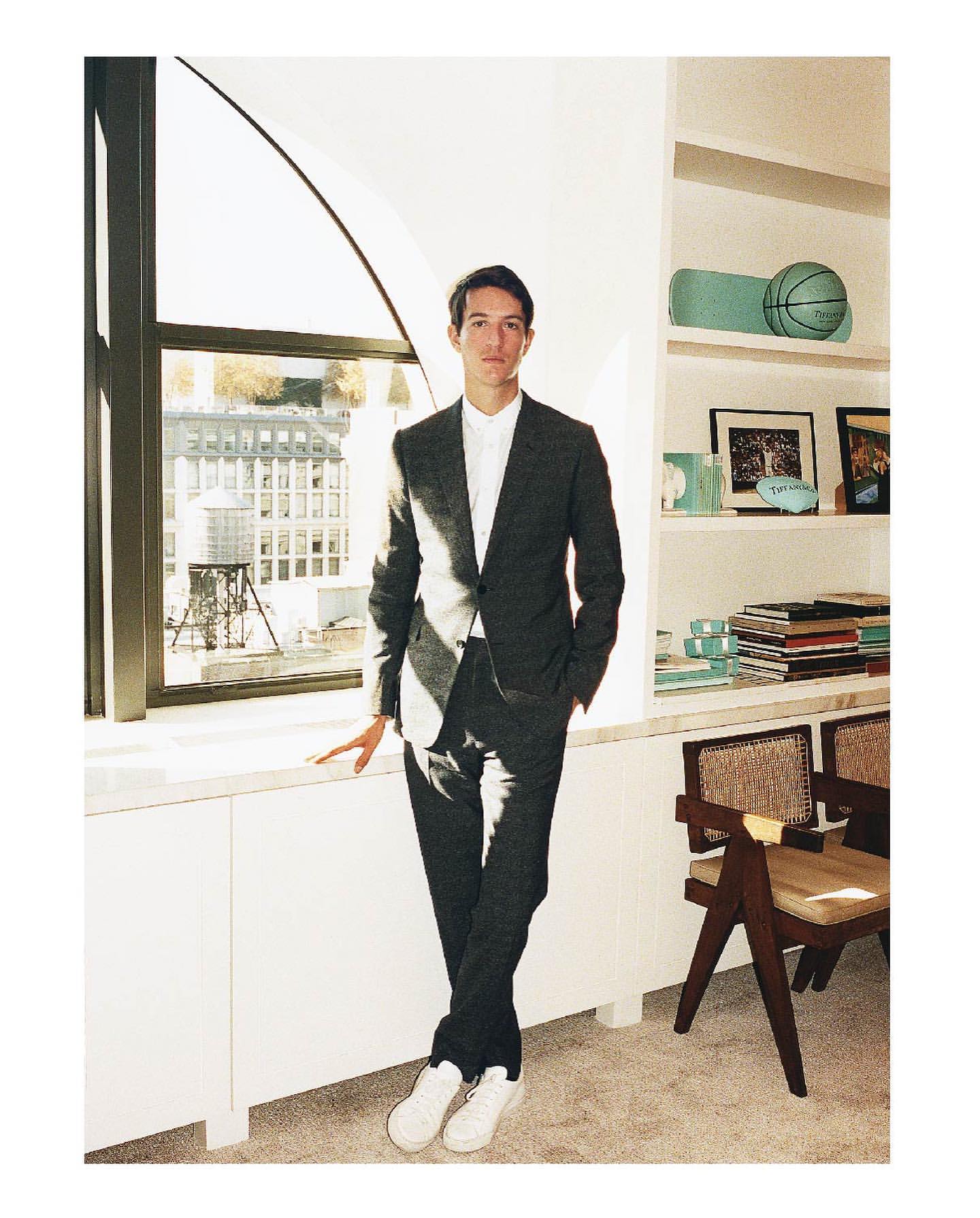 Well-dressed man in suit standing by window with city view, styled with white sneakers and bookshelf in a bright indoor setting.