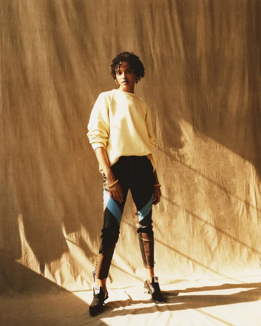 A slender young woman stands in front of a textured backdrop, wearing a pale yellow sweatshirt and athletic pants, with black shoes.