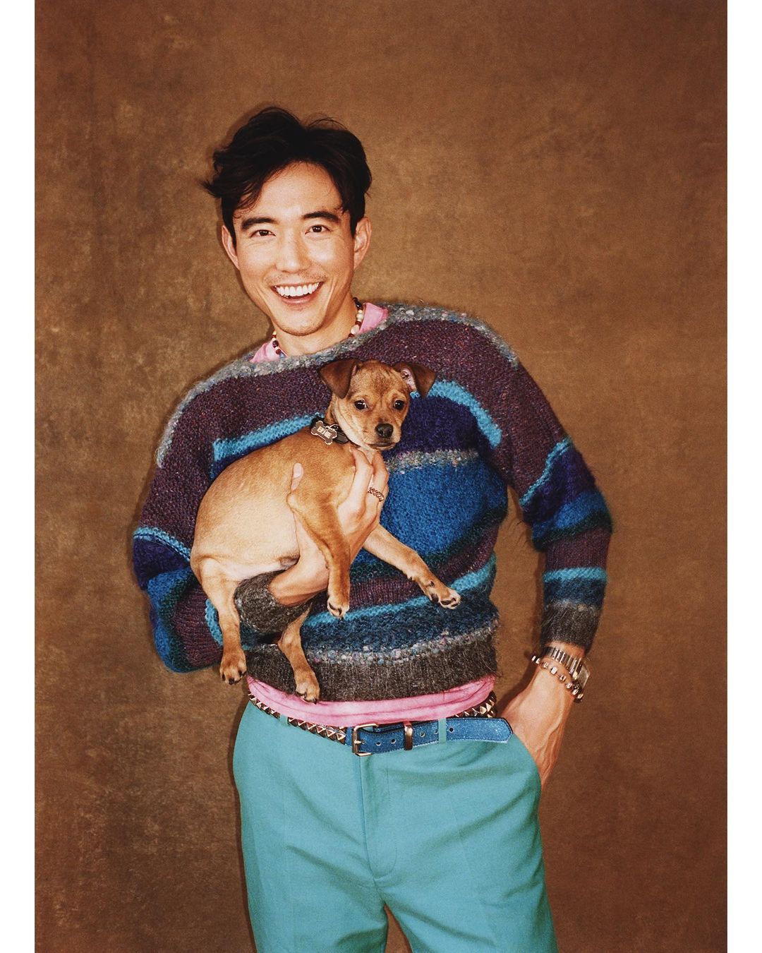 Adult male smiles and holds a dog. He wears a colorful sweater and blue pants, posing indoors against a brown textured backdrop.