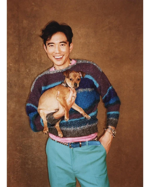 Adult male smiles and holds a dog. He wears a colorful sweater and blue pants, posing indoors against a brown textured backdrop.
