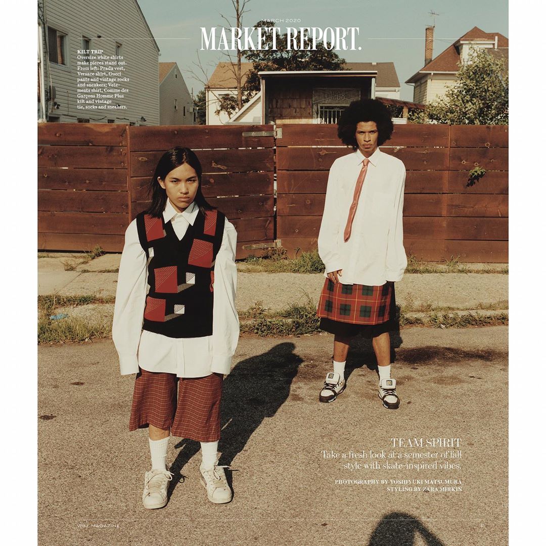 Two young people stand on a street wearing oversized shirts, kilts, and sneakers, with houses and a wooden fence in the background.