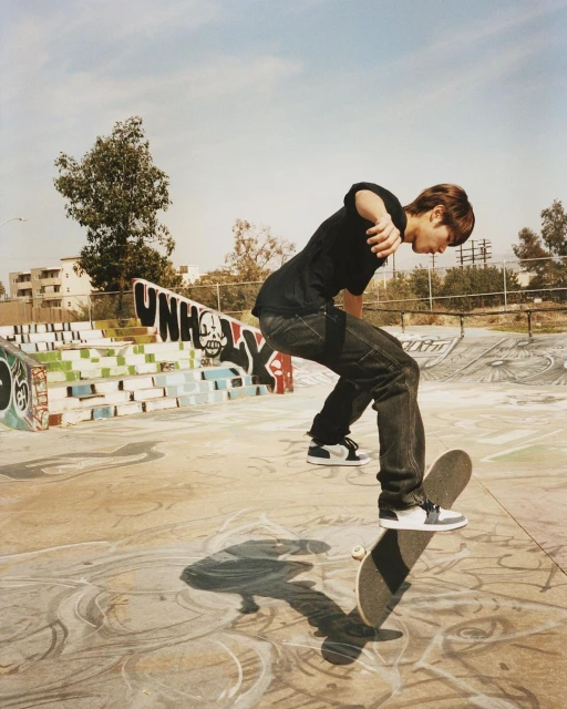 A skateboarder performs a trick at a skate park, wearing a black shirt and jeans, with graffiti and buildings in the background.