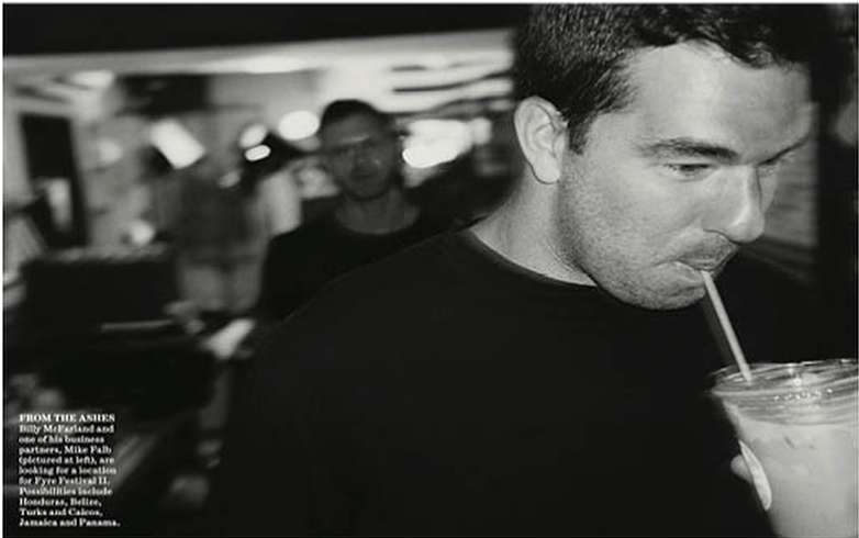 Black and white portrait of a man drinking from a plastic cup with a straw, with a blurred background of a second man indoors.