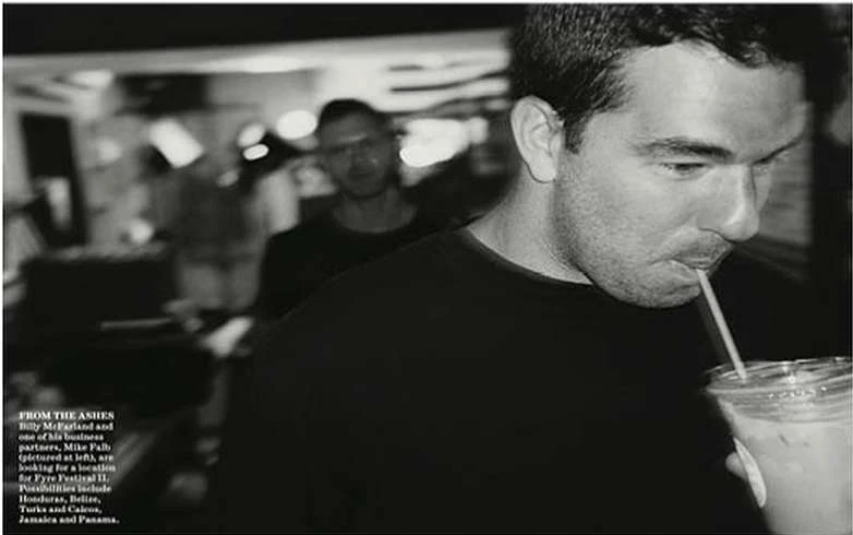Black and white portrait of a man drinking from a plastic cup with a straw, with a blurred background of a second man indoors.