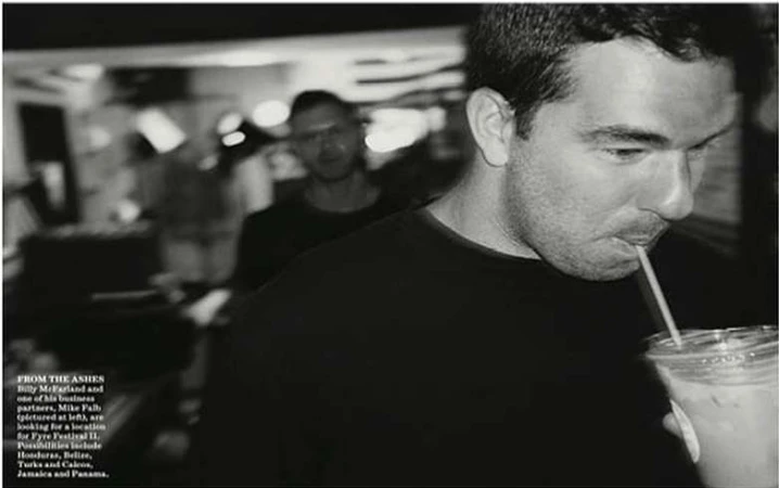 Black and white portrait of a man drinking from a plastic cup with a straw, with a blurred background of a second man indoors.