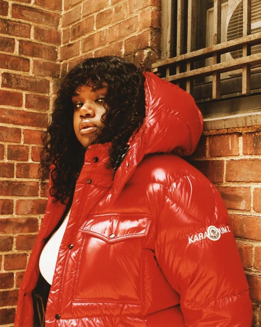 An adult female wearing a red puffer coat poses in front of a brick wall with a barred window, exhibiting a serious facial expression.
