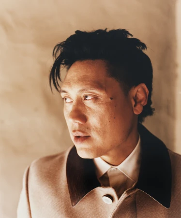 Portrait of an Asian man looking away, wearing a tan coat with dark lapels over a white collared shirt against a neutral wall.
