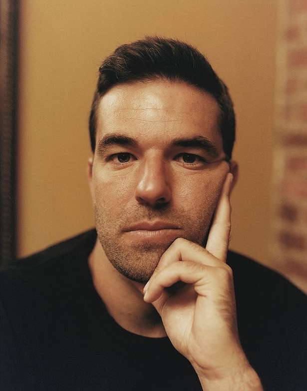 Close-up portrait of an adult male with short dark hair, resting his chin on his hand. Shot in a dimly lit indoor environment.