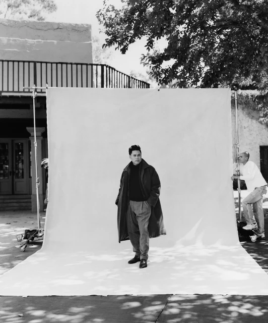 Black and white photo of an adult man posing in front of a white backdrop, wearing a dark coat and trousers, outdoors during daytime.