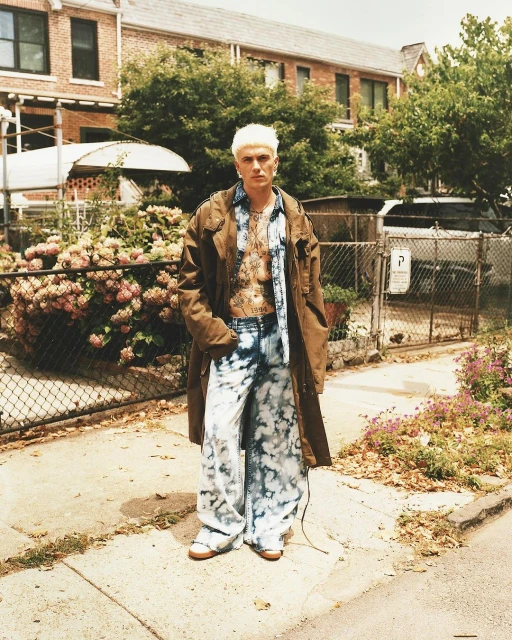 A person with bleached blonde hair wearing a brown coat, blue shirt, and blue and white tie-dye pants, stands on a sidewalk in front of a building.