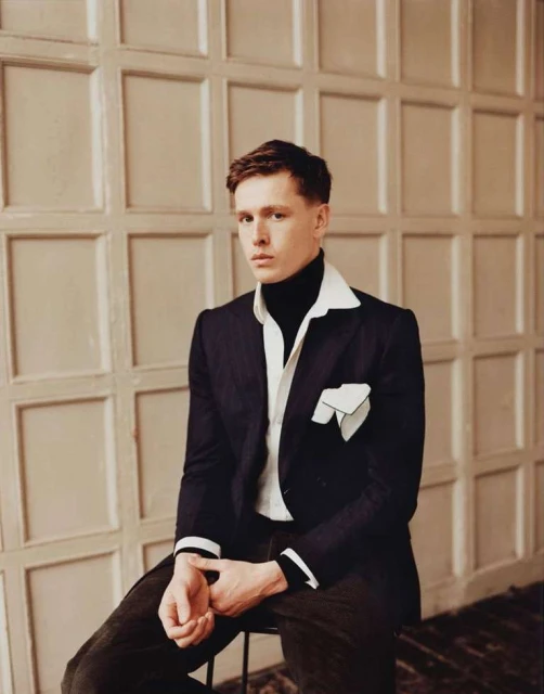 Portrait of a young adult male seated in a photo studio, wearing a black suit and white dress shirt with dark pants in a neutral setting.