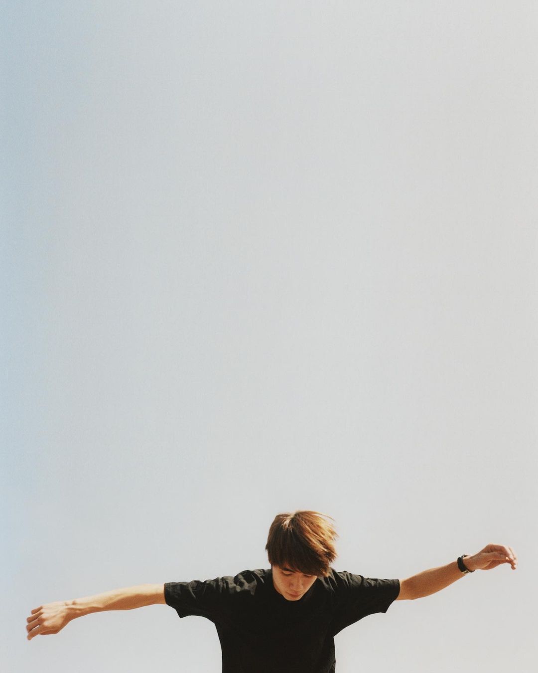 Adult male with outstretched arms against a light blue sky. Wearing a black t-shirt and a wrist watch. Shot from a low angle.