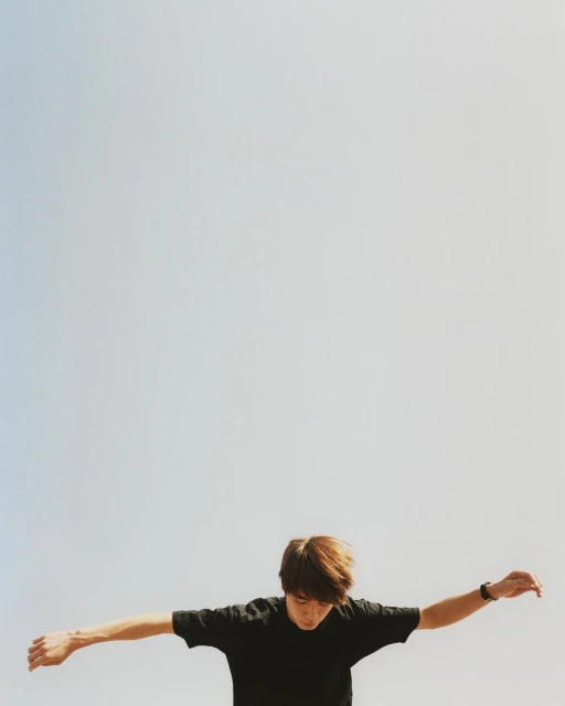 Adult male with outstretched arms against a light blue sky. Wearing a black t-shirt and a wrist watch. Shot from a low angle.