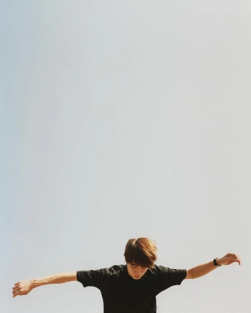 Adult male with outstretched arms against a light blue sky. Wearing a black t-shirt and a wrist watch. Shot from a low angle.