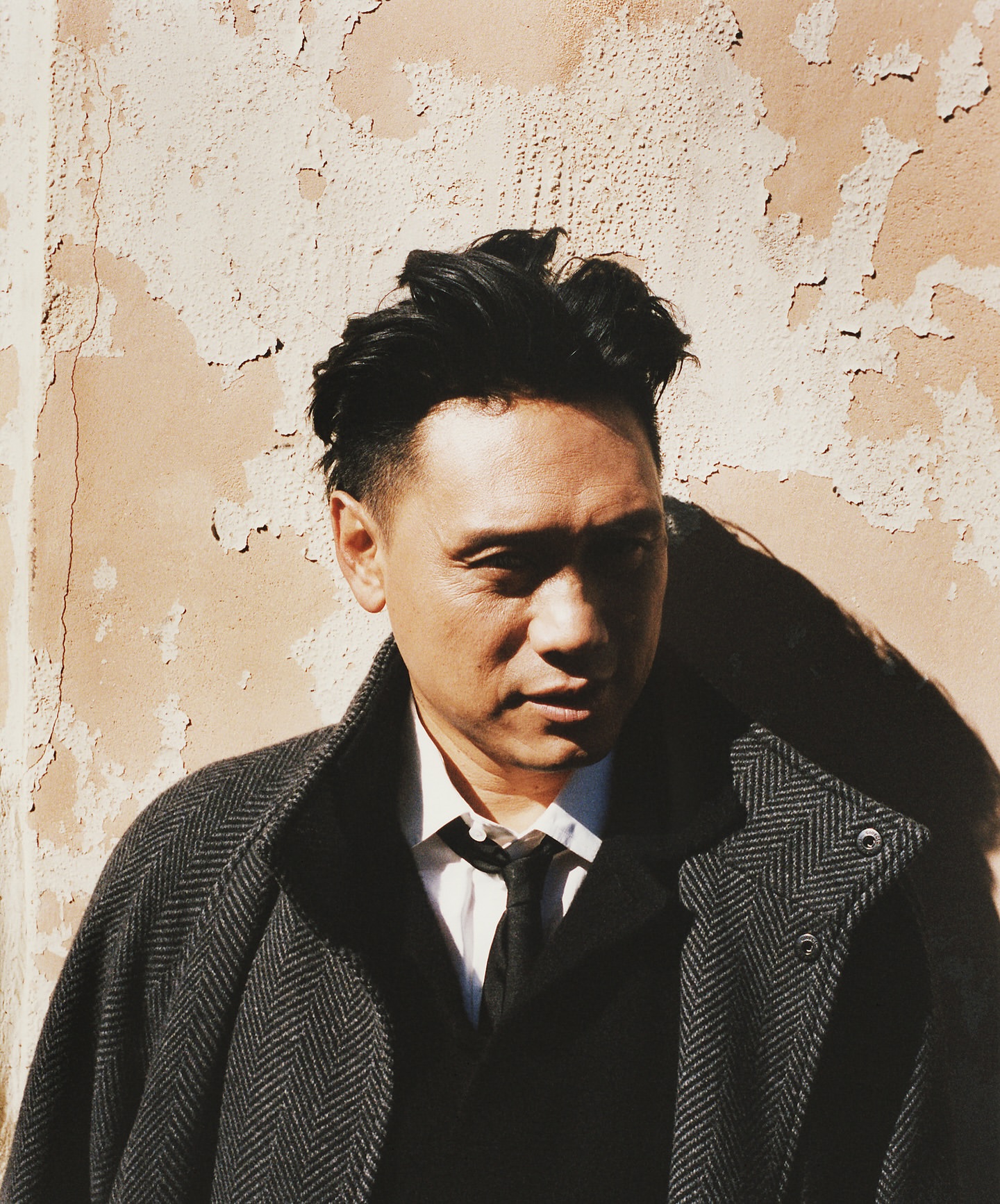 Headshot of a man with black hair in a herringbone overcoat standing in front of a weathered peach-colored wall, outdoor portrait.