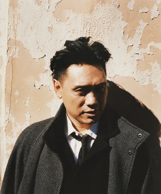Headshot of a man with black hair in a herringbone overcoat standing in front of a weathered peach-colored wall, outdoor portrait.