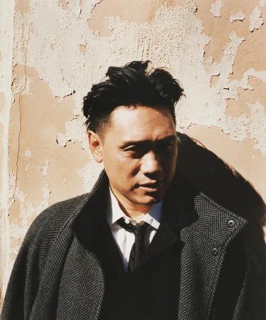 Headshot of a man with black hair in a herringbone overcoat standing in front of a weathered peach-colored wall, outdoor portrait.