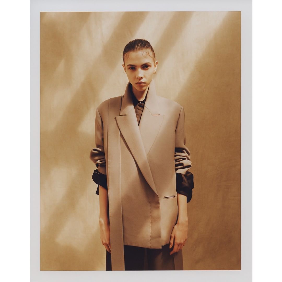 Young woman in an oversized beige blazer and black shirt against a soft brown backdrop with cast shadow stripes in a studio shot.