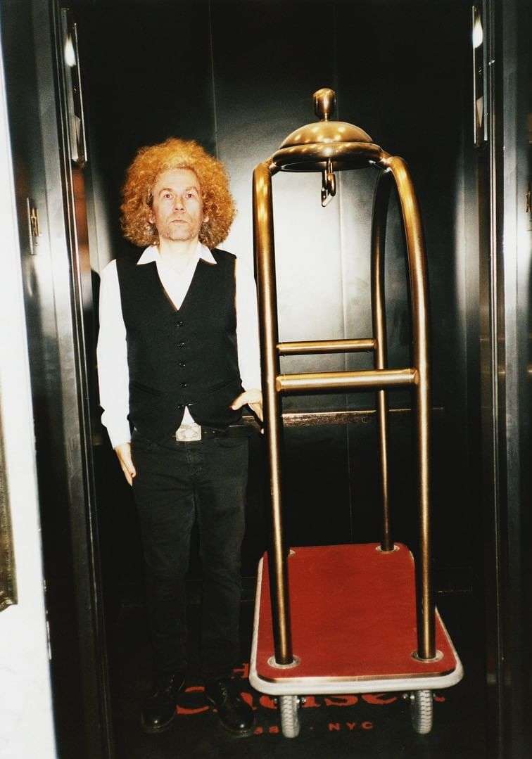 Adult male with curly red hair stands next to a brass luggage cart in a modern elevator with gray walls and a dark-colored floor.