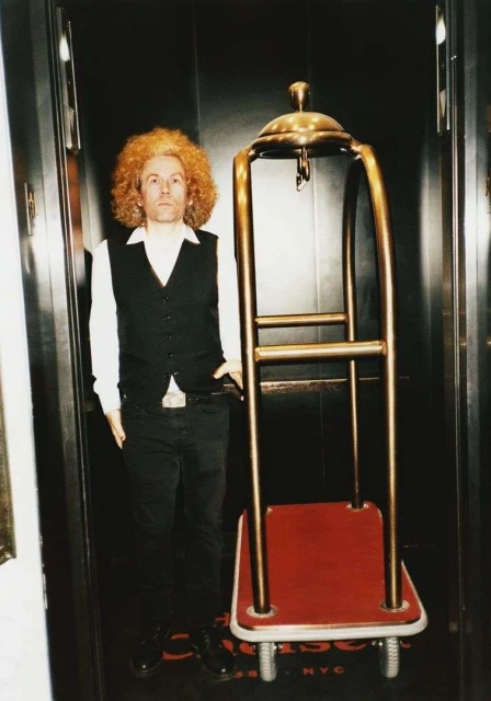Adult male with curly red hair stands next to a brass luggage cart in a modern elevator with gray walls and a dark-colored floor.