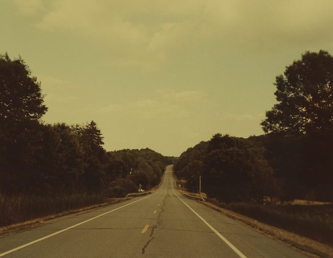 Straight asphalt road stretches into the distance, framed by green trees and a light sky on a clear day, promoting a peaceful scene.
