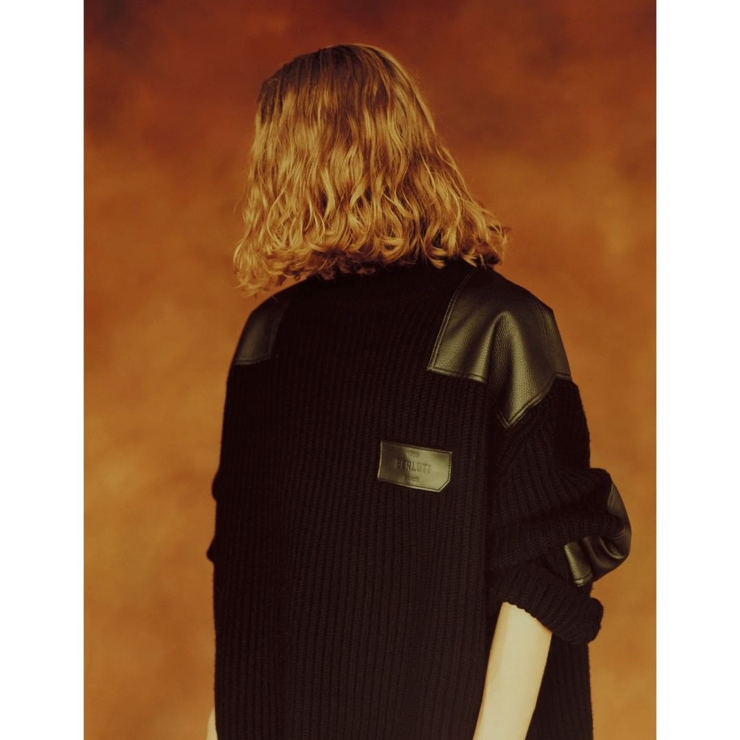 Back view of adult with wavy blonde hair wearing a black knit sweater with leather shoulder patches against an orange background.