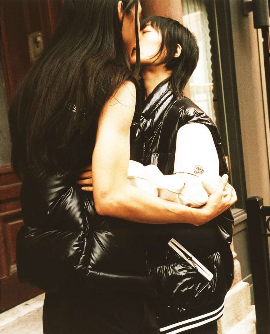 Two adults kiss while embracing outside in black puffy jackets. The couple stands close together next to a building with light filtering through.