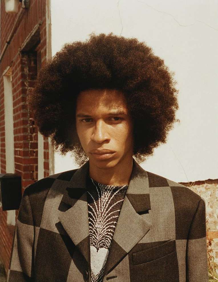 Portrait of a young adult male with an Afro hairstyle, wearing a patterned sweater and checkered blazer, standing against a brick wall.