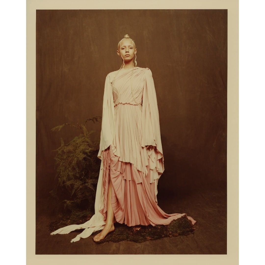 A woman in a long, flowing light pink gown with an off-the-shoulder design, posing in a studio with mossy ground cover and lush greenery.