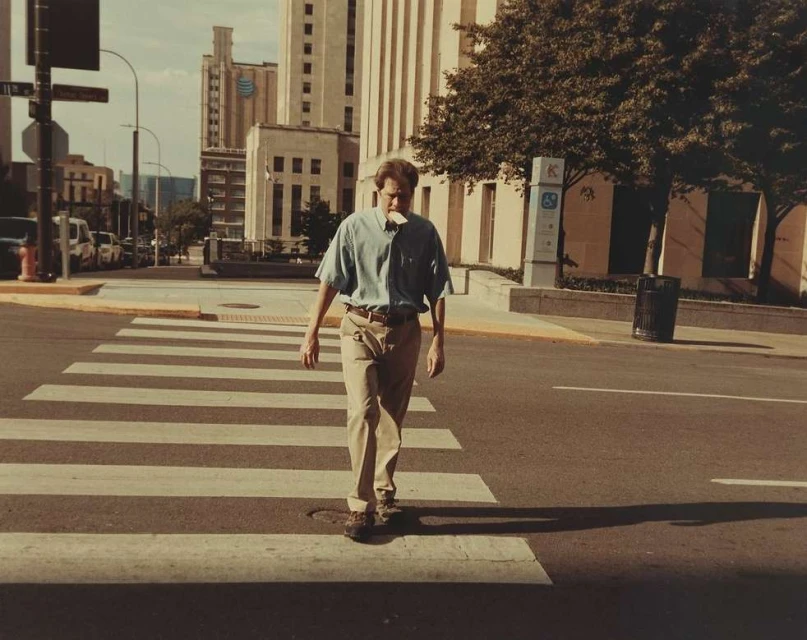 Adult male crosses a pedestrian walkway in an urban setting with buildings and trees in the background on a sunny day.