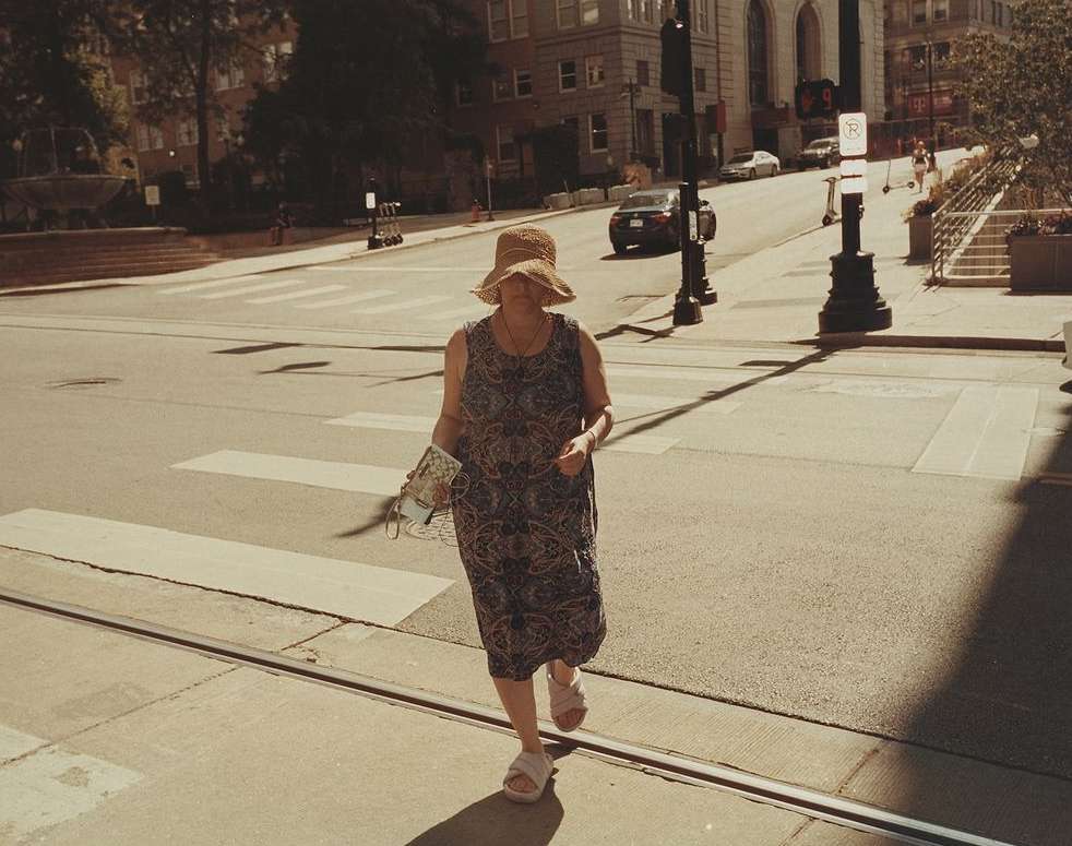 Woman in sunhat crossing street on crosswalk in summer with cars and city buildings in the background in an urban environment.