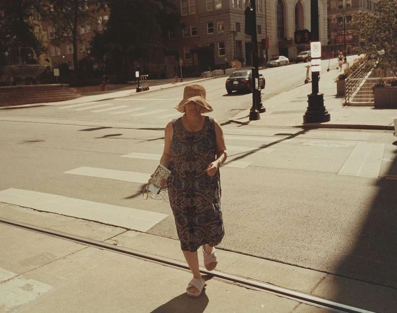 Woman in sunhat crossing street on crosswalk in summer with cars and city buildings in the background in an urban environment.