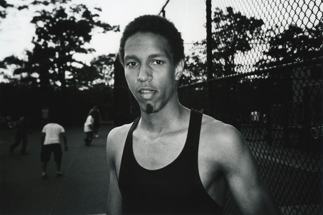 Black and white portrait of a young man in a dark tank top outdoors near a chain link fence by Yoshiyuki Matsumura