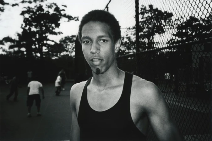 Black and white portrait of a young man in a dark tank top outdoors near a chain link fence by Yoshiyuki Matsumura