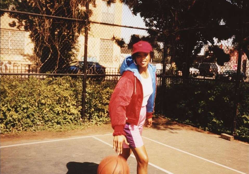 A woman dribbles a basketball on an outdoor court. She wears a red cap, red and blue jacket, and pink shorts in the sunny day.