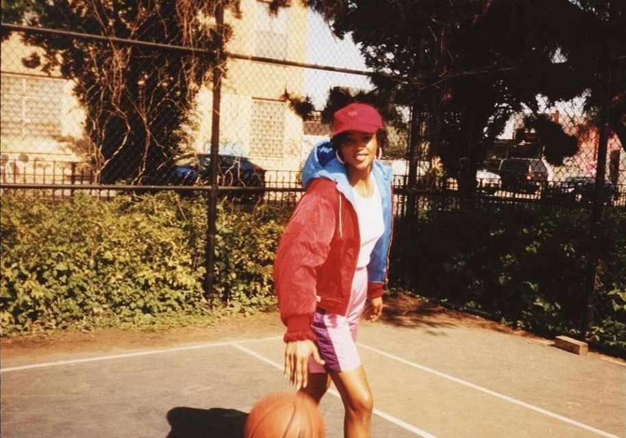 A woman dribbles a basketball on an outdoor court. She wears a red cap, red and blue jacket, and pink shorts in the sunny day.