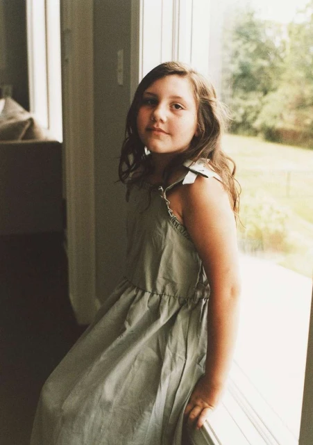 Young girl in light green dress sitting by the window with natural light, looking at the camera with a neutral facial expression.