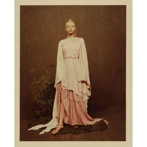 Model with blonde hair standing barefoot in a pleated cream and pink gown before a dark textured wall and foliage.