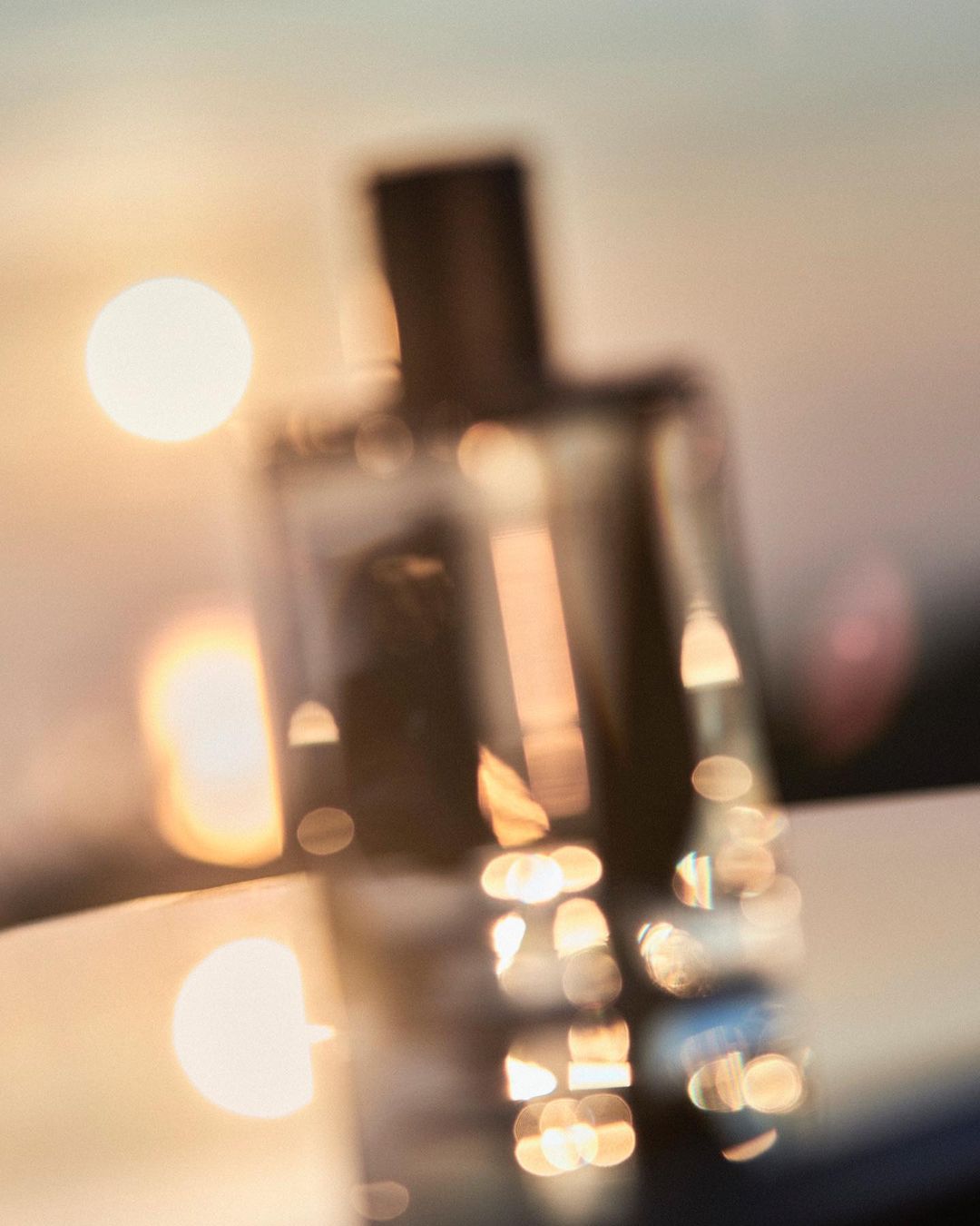 Close-up of a blurred perfume bottle on a reflective surface with a golden sunset background, creating a warm, luxurious atmosphere.