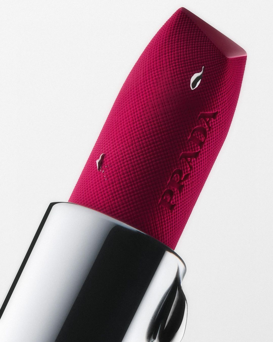 Close-up macro shot of a vibrant pink Prada lipstick with textured surface in a silver case against a white background