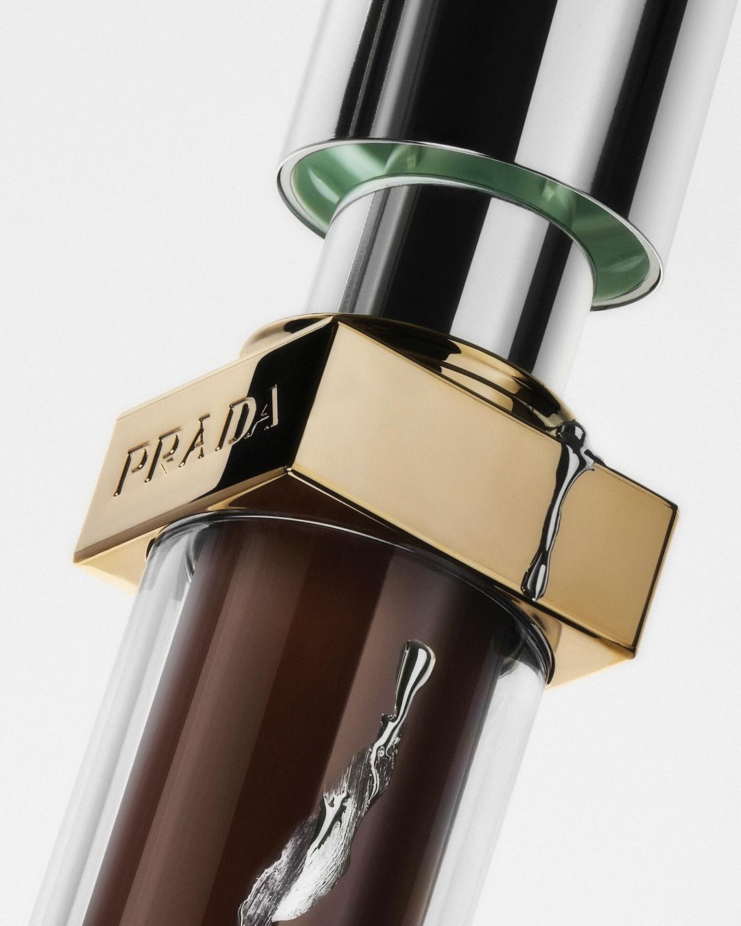 Close-up of a Prada lip gloss featuring a clear tube, a golden top with the Prada logo, and brown gloss.