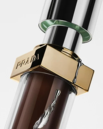 Close-up of a Prada lip gloss featuring a clear tube, a golden top with the Prada logo, and brown gloss.