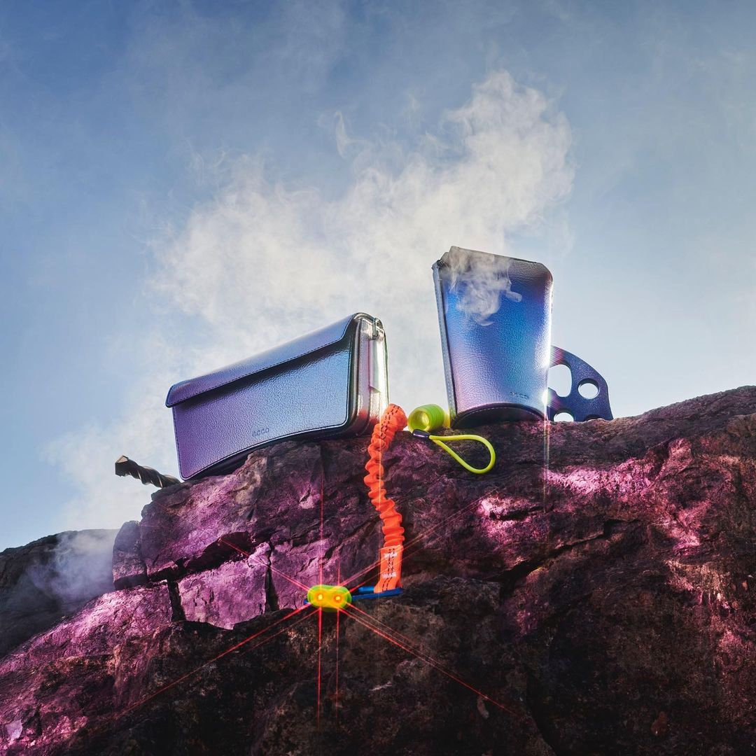 Two metallic-finish containers and an orange bungee cord sit on a rocky surface against a blue, cloudy sky in a product shot.