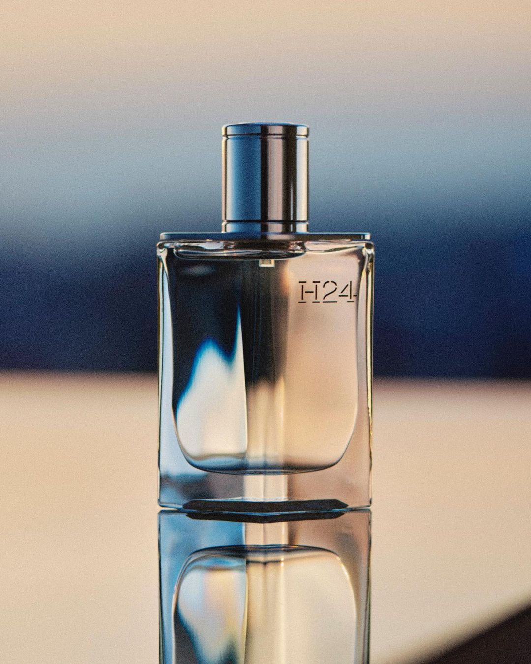 Elegant square-shaped perfume bottle with a metallic cap and H24 lettering, set against a blurred blue background and reflective surface.